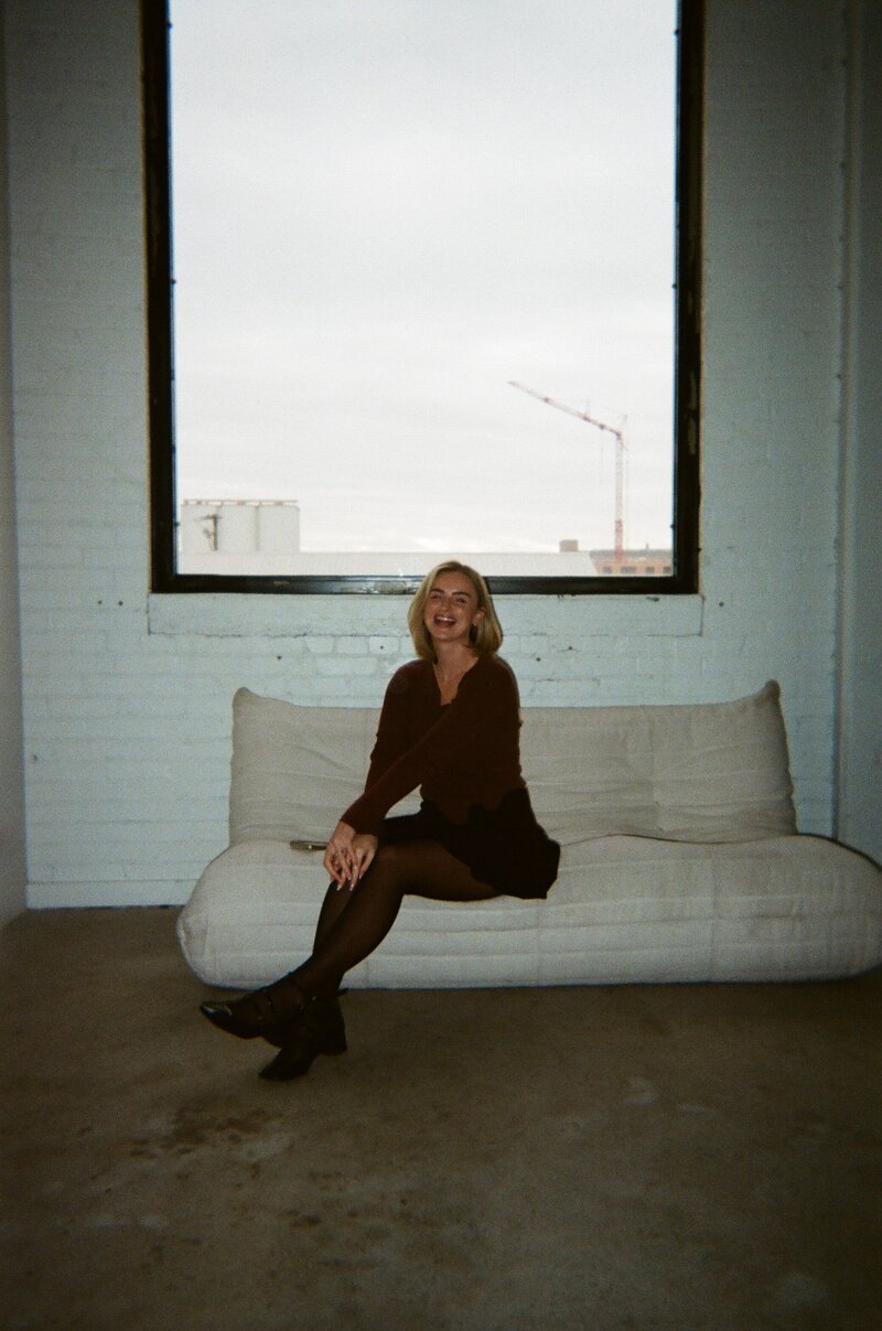 Jaiden sitting on a white couch in a studio with a big window behind her