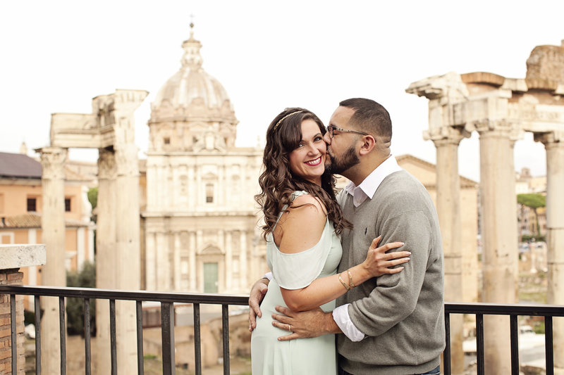 A woman being kissed on the cheek in front of the Roman Forum. Taken by Rome Honeymoon Photographer, Tricia Anne Photography
