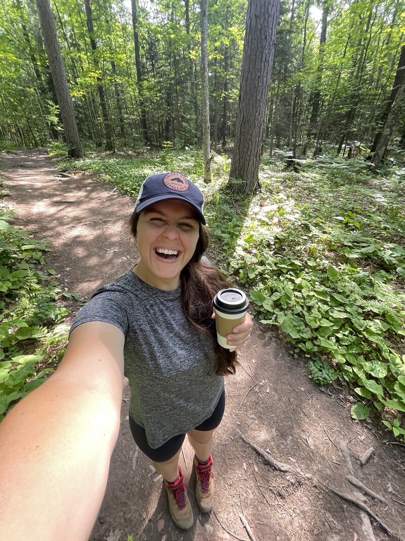 Morgan, a wedding photographer in Duluth, MN, smiling for a selfie while holding coffee and wearing hiking boots and a hat from Cornucopia, WI, during a hike