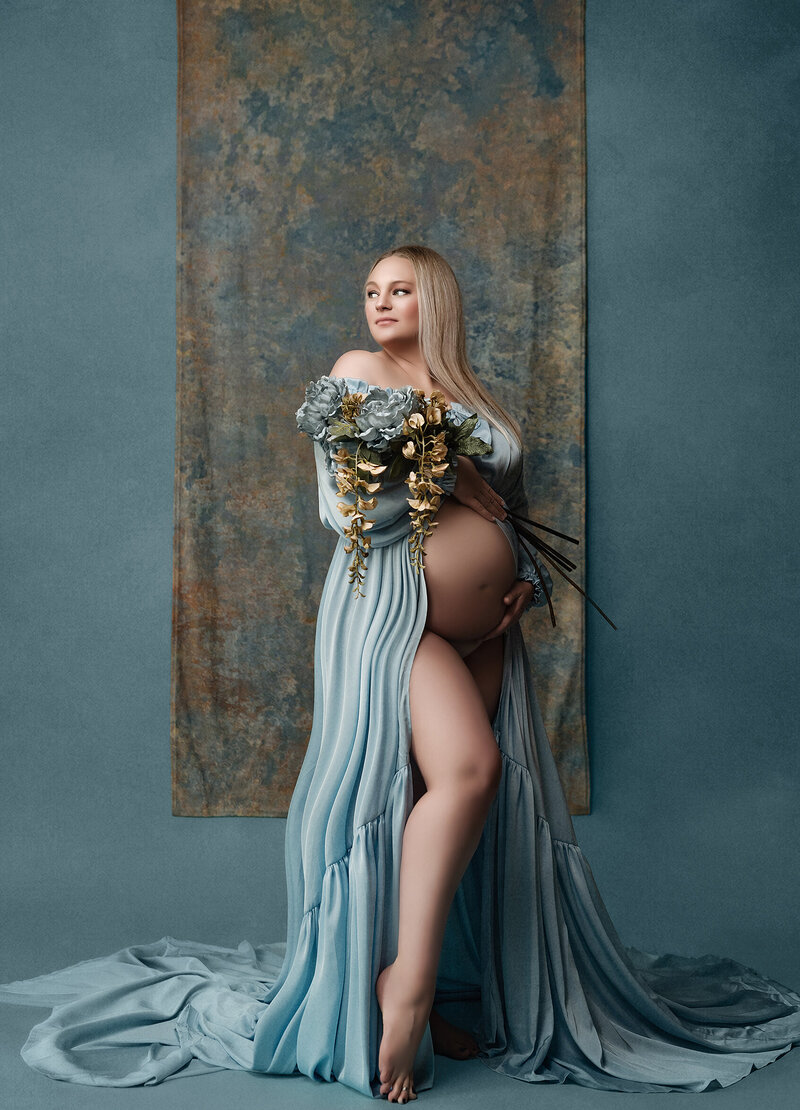 standing posed mom to be holding flowers and belly on the shade of blue in studio