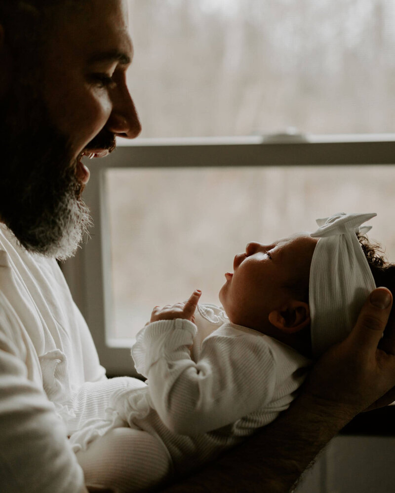 New dad smiling at his newborn baby girl and holding her in front of a window in their home.