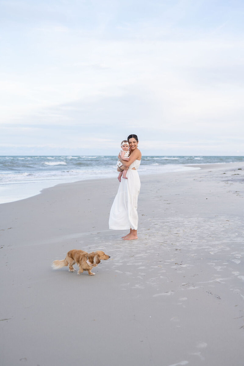 mother wearing a long white dress standing on the beach holding a baby girl as their family dog walks by