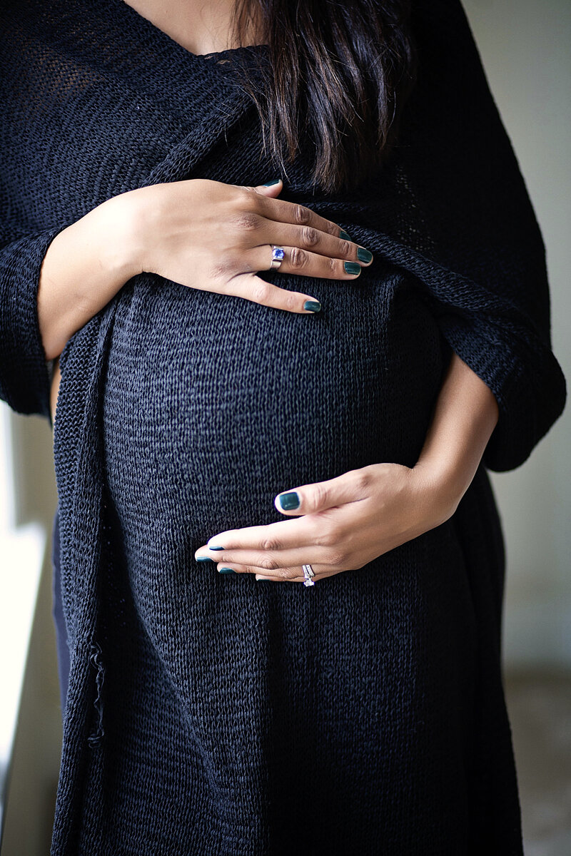 New mom wrapped in black sweater standing by the window holding her belly