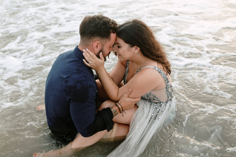 A couple sits in the shallow waves of the ocean, sharing an intimate moment