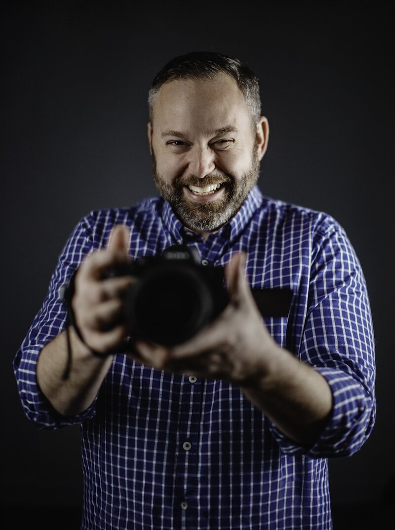 A Columbus wedding photographer captures a moment with a beaming smile, holding a professional camera. Dressed in a blue and white checked shirt, his expression is full of joy and enthusiasm, illustrating his passion for photography. The dark, neutral background emphasizes his cheerful demeanor and professional attire, perfect for a wedding photographer who brings both skill and spirit to his work.