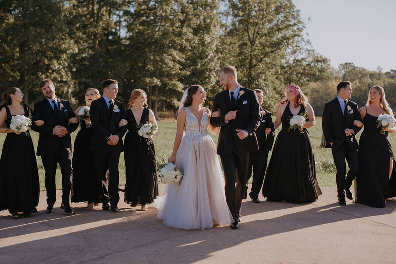 A bride and groom walk arm in arm, smiling at each other. They're accompanied by a joyful wedding party dressed in black attire, holding bouquets. This serene scene unfolds at an outdoor wedding venue in Virginia on a sunny day, with trees beautifully framing the moment.