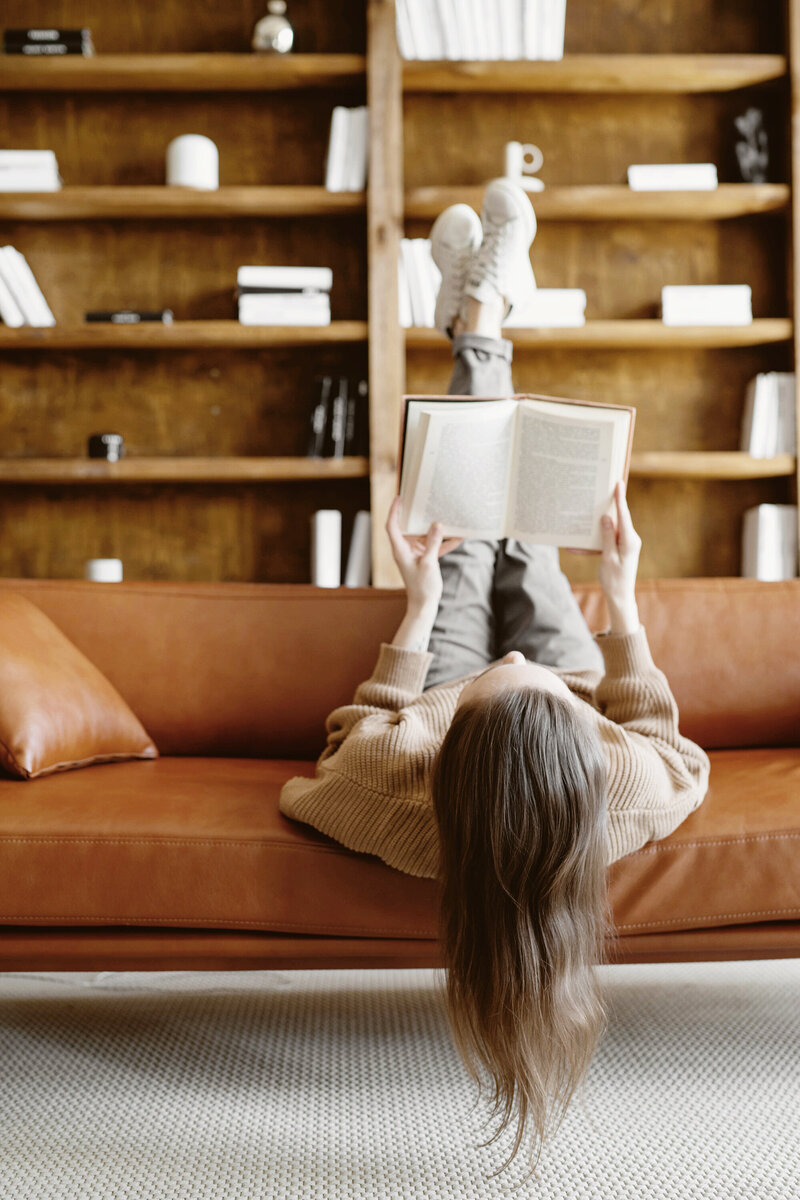 Catherine Shuman lies upside down on a caramel leather sofa reading a book, legs up on the backrest, with long hair cascading over the cushion and wooden bookshelves in the background. A sidebar reveals personal favorites like coffee + Stanley, exploring Alaska, Dove Cameron playlists, a raspberry chocolate martini, and INTJ — showcasing the human side of an operations consultant for women-led architecture and interior design firms.