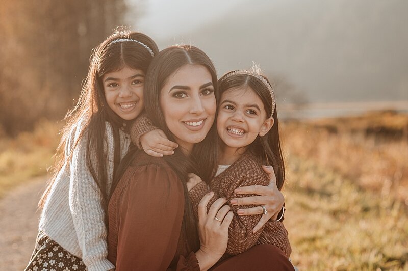 Two happy toddler sisters smile and hug their happy mom in a park trail at sunset