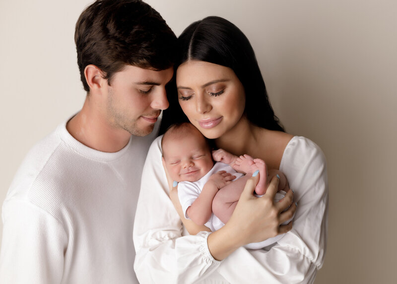 Studio newborn image. Mom is holding baby and Dad is standing behind mom, their heads touching. Mom and Dad's eyes are closed and have a closed-mouth smile. Baby's eyes are closed and baby is smiling.