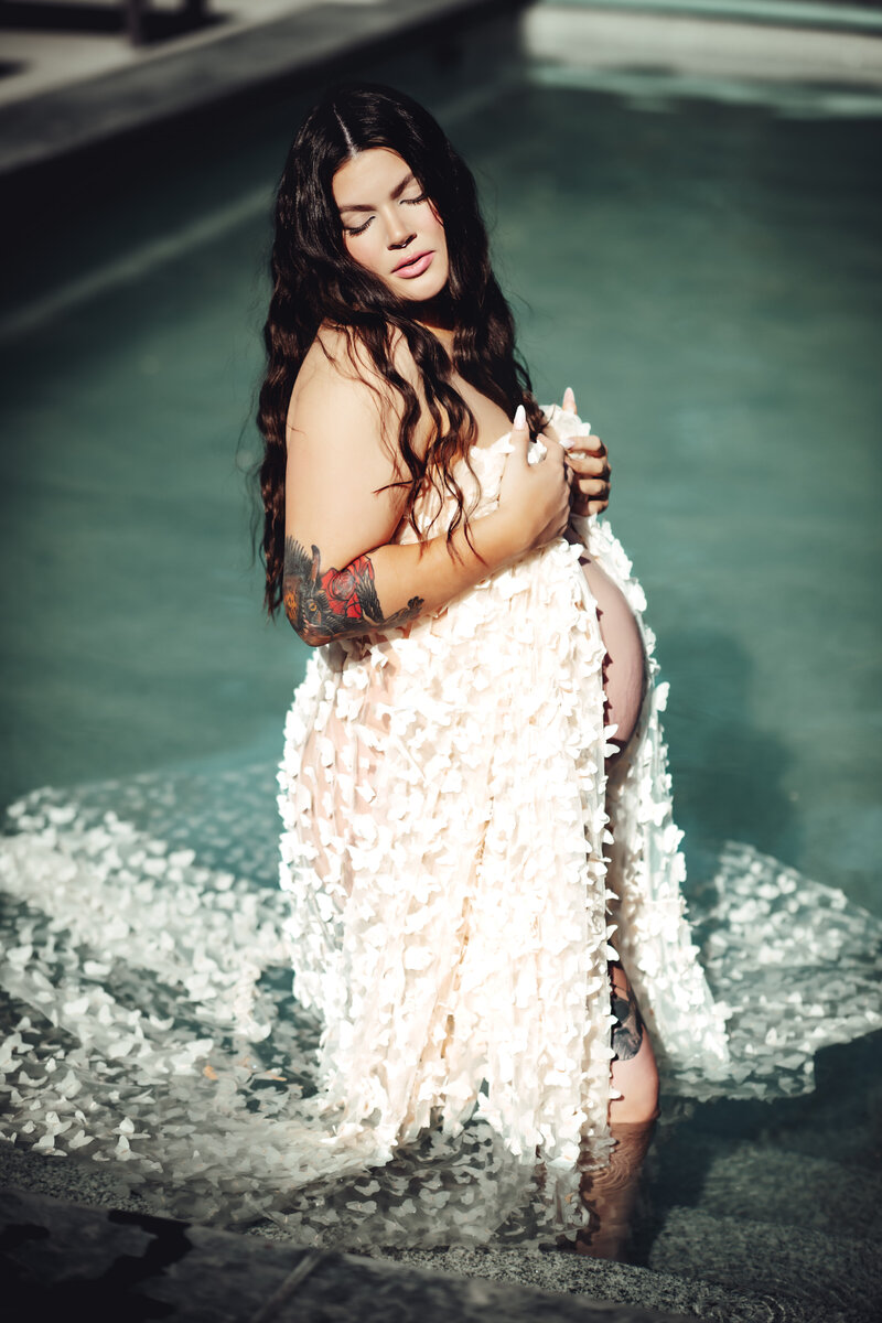 Gorgeous pregnant woman draped in sheer white fabric standing in a pool. 