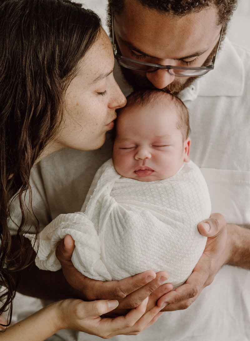 Newborn photo of dad and baby wearing white while holding baby and mom and dad kissing baby's head in studio in Annapolis Maryland photographed by Bethany Simms Photography