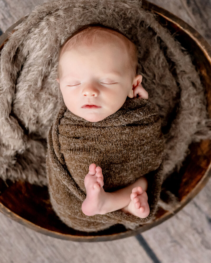 A newborn baby in a brown swaddle sleeps in a wooden bucket