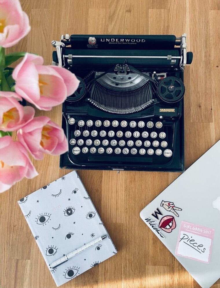 Martina Menzini's desk featuring a laptop, vintage typewriter, notebook, pen, and vase of flowers, showcasing inspiration for her 'Pieces' column.