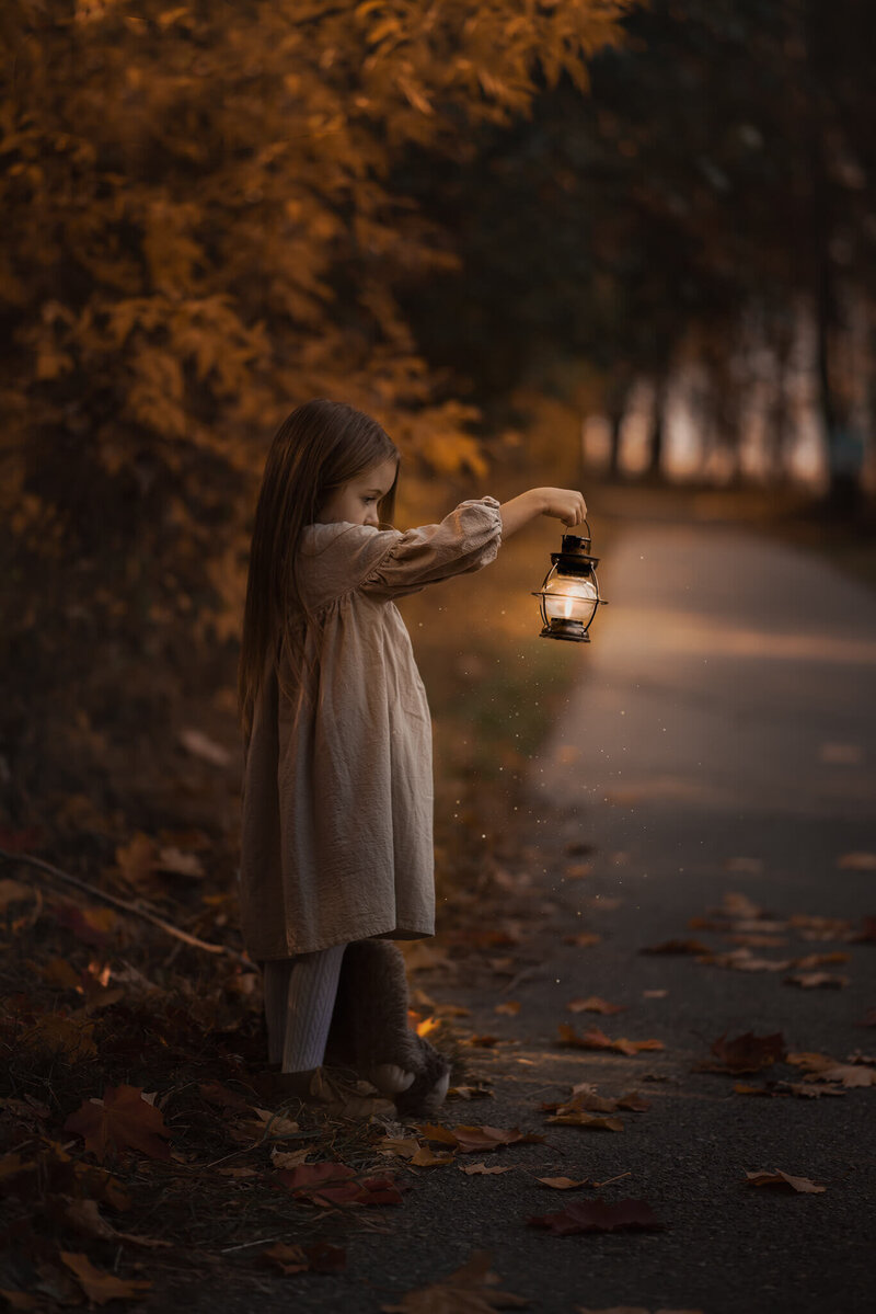 Along a pathway in a park stands a little girl with brown hair in a dress holding up a glowing lantern while looking into it. Photo taken by Brittany Danielle