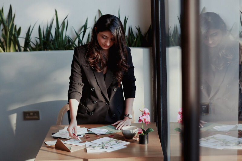 Komal, a talented artist and designer, works on her latest project in a cafe, dressed in a stylish black blazer. Specializing in bespoke wedding stationery and watercolor illustrations, she crafts personalized designs with precision. Based in Oman, Komal’s art reaches clients worldwide.
