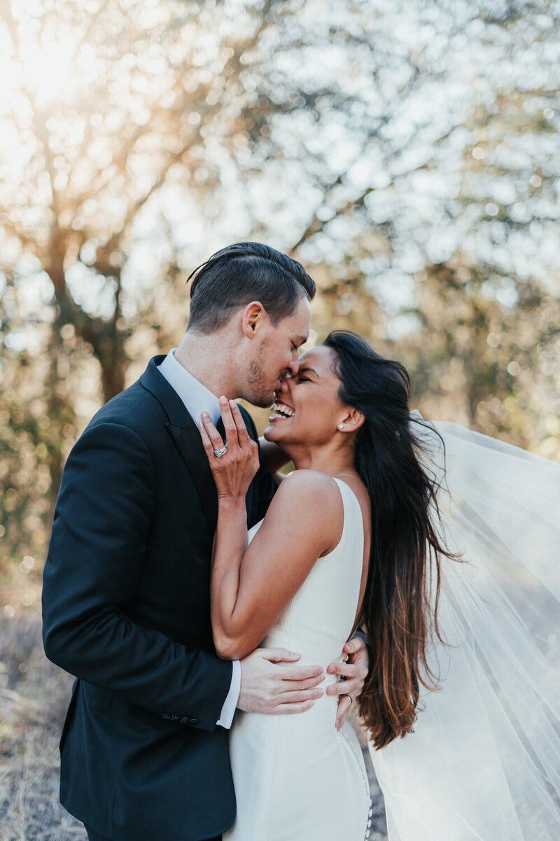 Groom embraces beautiful dark-haired bride and leans in for a kiss, while she laughs and smiles. The wind is blowing her dark hair and veil. Photo taken by Orlando Wedding Photographer Four Loves Photo and Film.