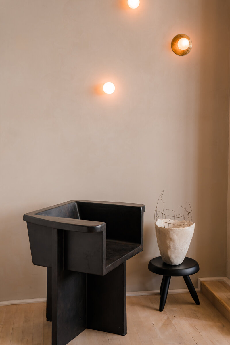 Stylish and sculpted black wooden chair with asymmetrical legs standing next to a black mini stool and decorative beige vase