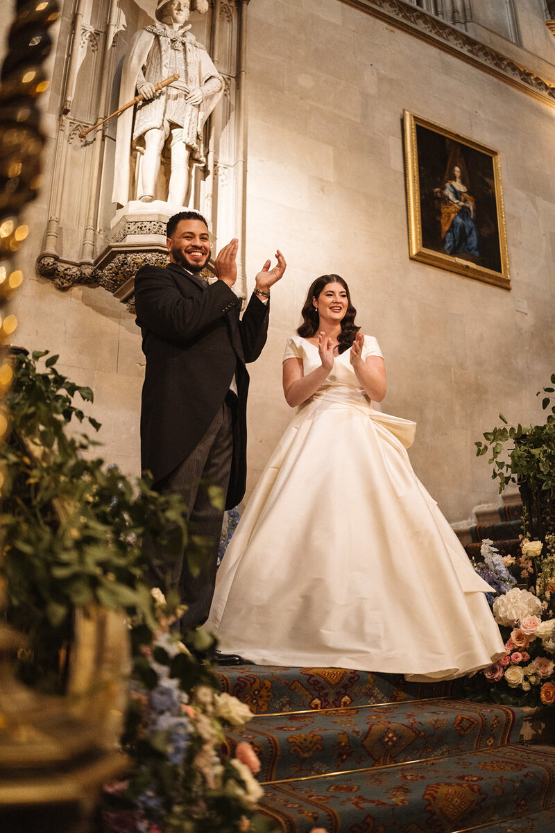 A joyful American couple stands on the grand staircase of Ashridge House in Hertfordshire, clapping their hands in celebration. The bride, radiant in a flowing white gown, and the groom, dapper in a tailored suit, share a moment of pure happiness against the backdrop of the historic venue’s ornate stonework. The soft glow of natural light highlights the intricate details of the staircase, adding to the magical atmosphere of their wedding day.