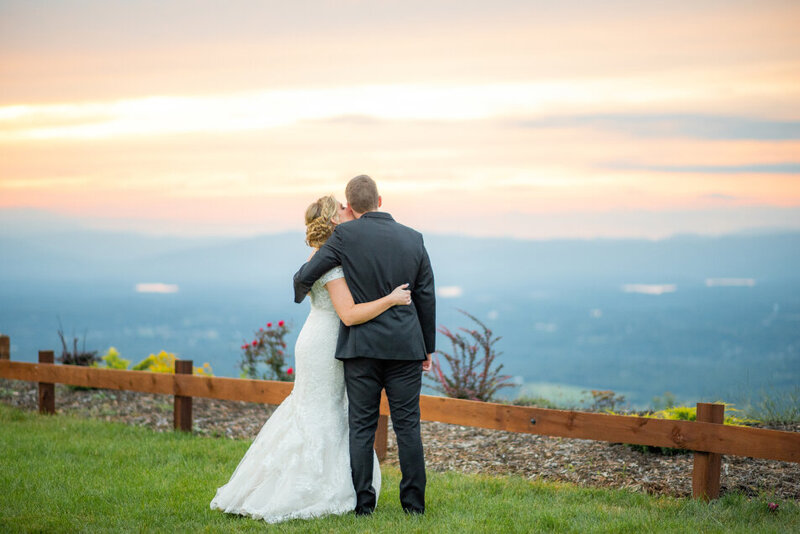 A bride and groom looking at the Blue Ridge Mountains at Hidden Hill Venue in Morganton NC. The bride is kissing the groom on the cheek.
