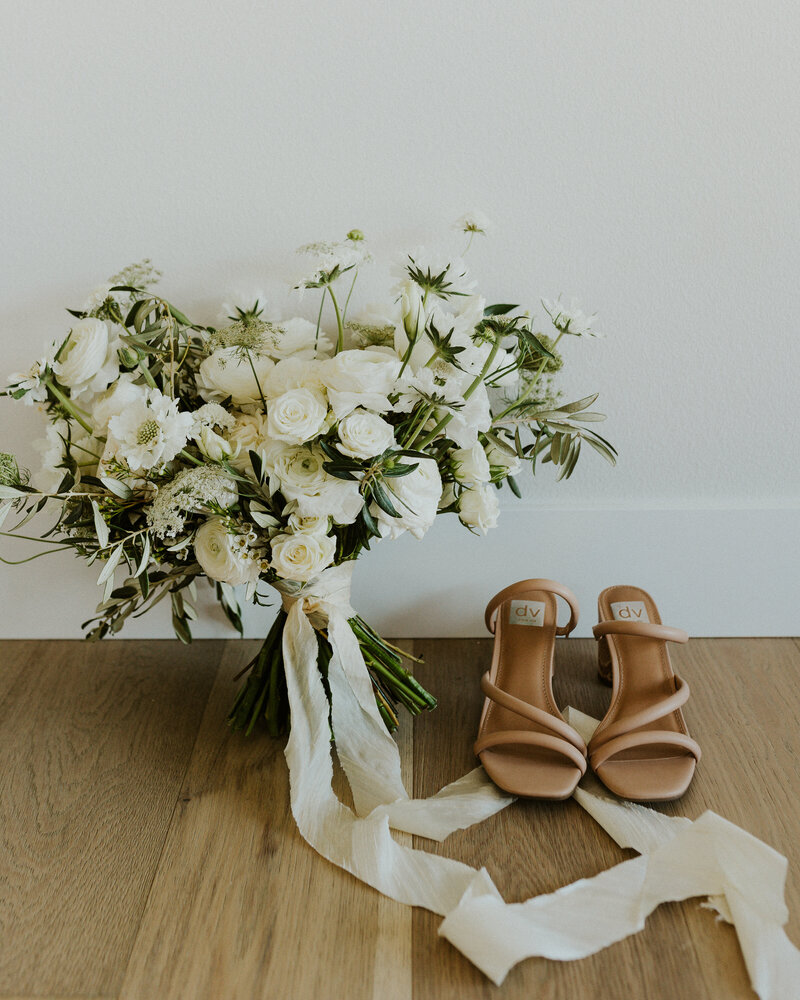 Light brown strappy heels sitting next to a white and green floral bouquet rapped with a cream ribbon