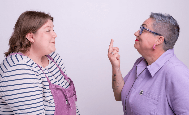 Pippa on the right, in a lilac jumpsuit, smiles and playfully holds a middle finger up at Lucy, who’s wearing a striped top and purple dungarees, during a Disobedient Business® Co. photoshoot.