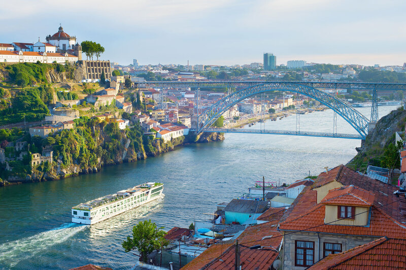 A scenic view of Porto, Portugal, featuring the iconic Dom Luís I Bridge over the Douro River. A boat is gliding along the river, with hillside buildings and a clear blue sky in the background.