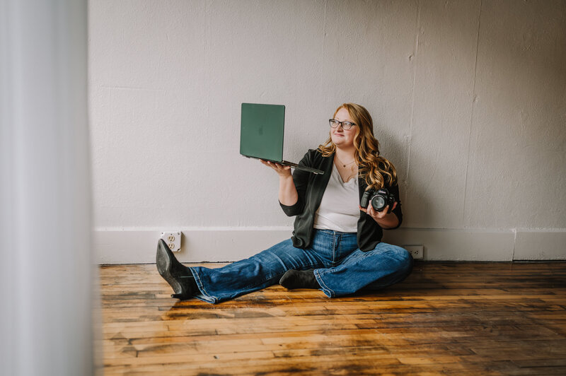 A women holding a laptop and camera sitting on the ground. 