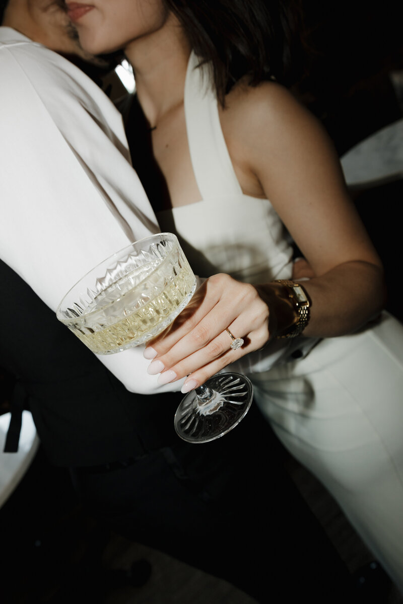 Bride holding champagne glass showing off her ring whiile her groom kisses her neck