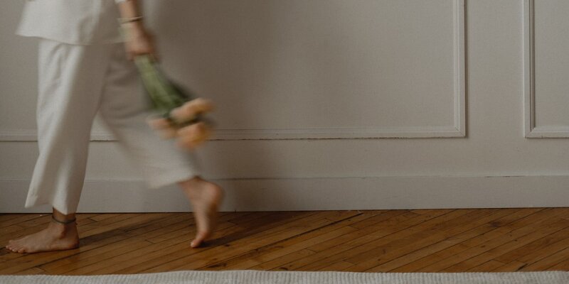 Soft-focus image of a woman in white pants walking barefoot on a wooden floor, holding a bouquet of flowers with a relaxed and chic vibe.