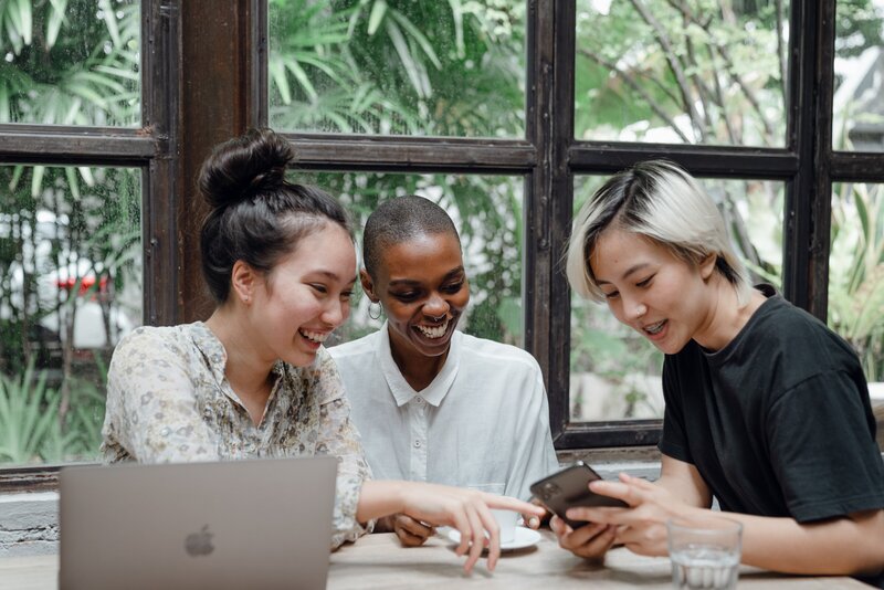 A group of 3 women from different racial backgrounds are sitting together. They appear to be working and looking at something on one of their phones and computer while smiling.