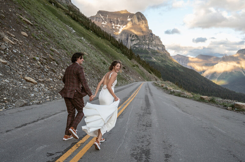 A newlywed couple walks down Going-to-the-Sun Road in Glacier National Park at golden hour, surrounded by rugged cliffs and glowing mountain light, the bride smiling back over her shoulder as Sydney Breann Photography captures their adventurous moment.