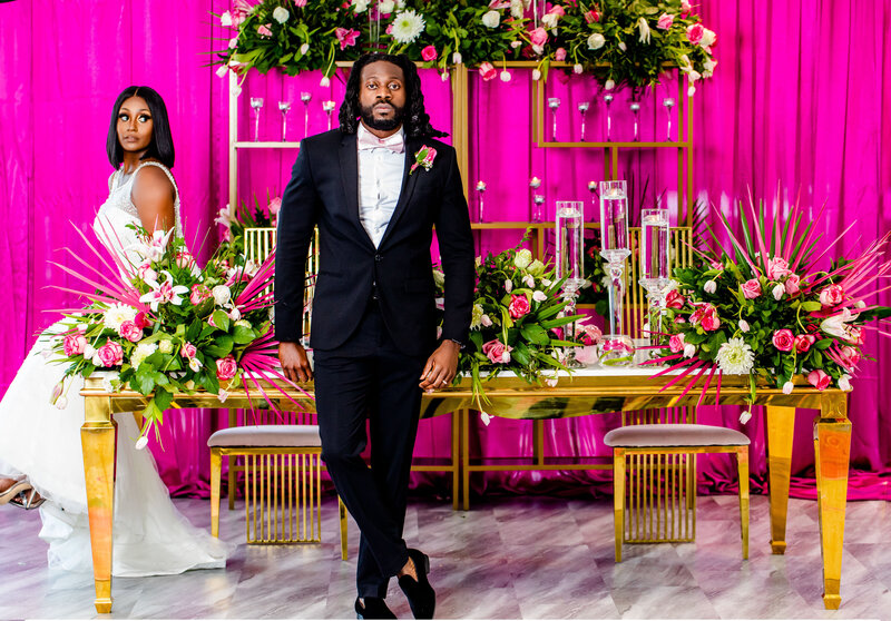 Celebrate love with this stunning image of a Black couple on their wedding day, featuring a gold estate table adorned with lush floral centerpieces. Designed by Beyond Expectations, the vibrant pink and green palm leaves create an enchanting atmosphere for their special day.