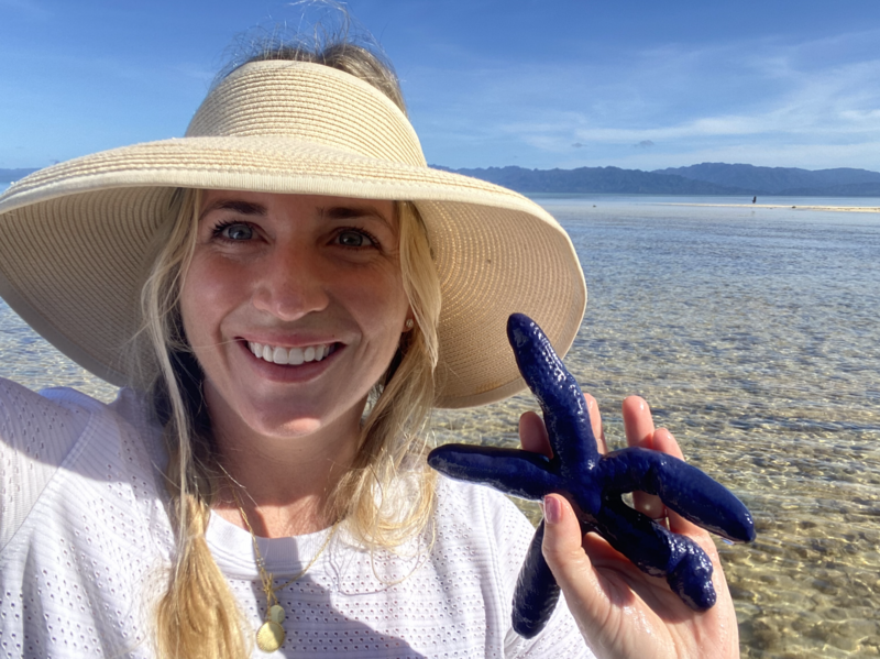 a woman in a hat holds a blue starfish while exploring tidal pools in Fiji.