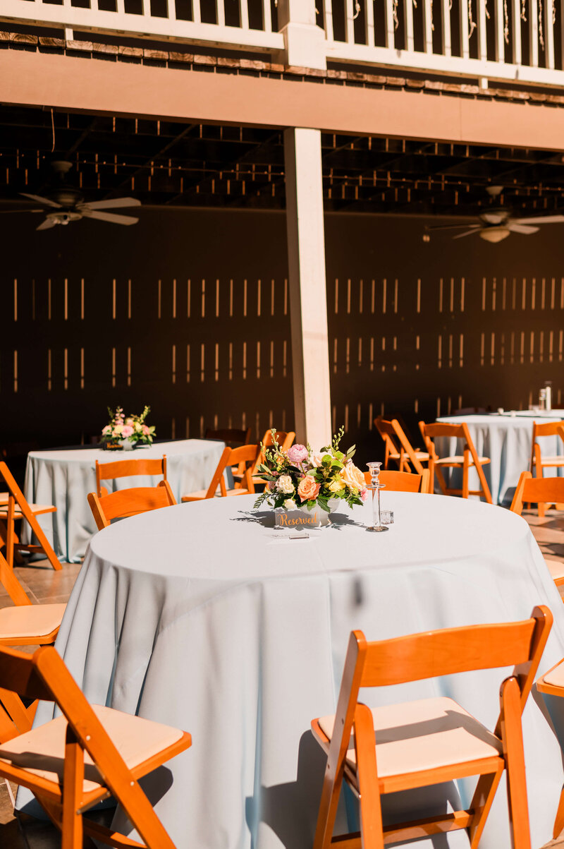 Round tables with light blue tablecloths and wooden chairs are set up outdoors. Each table has a floral centerpiece and place cards. Sunlight creates warm shadows on the scene.