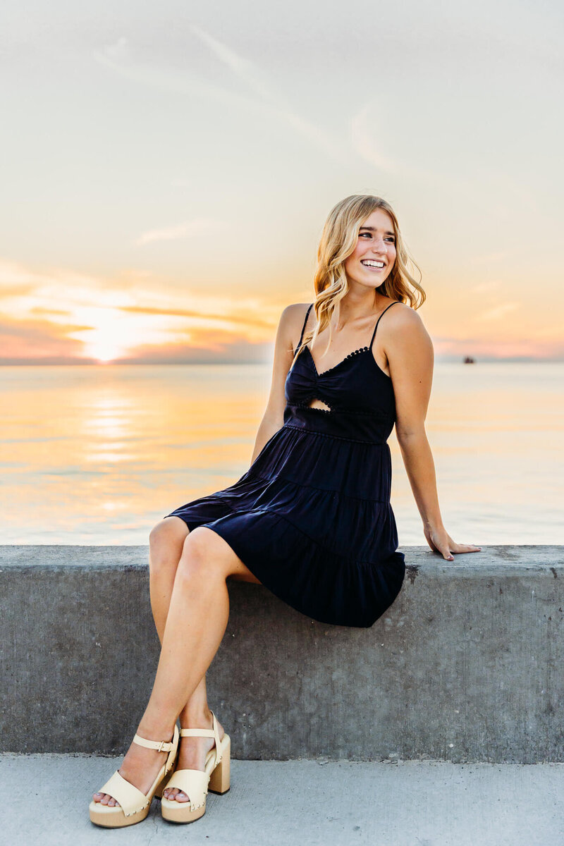 senior girl in blue dress sitting on a wall with the sunset behind her