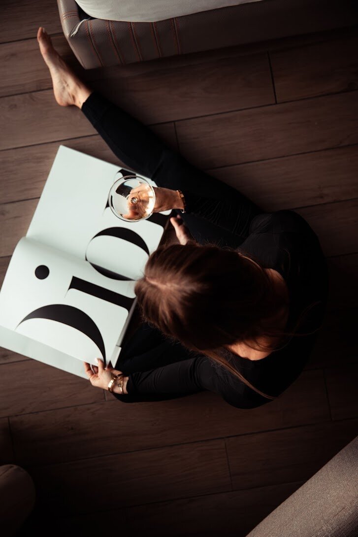 Woman sitting on the ground holding a glass of white wine and reading a Dior book