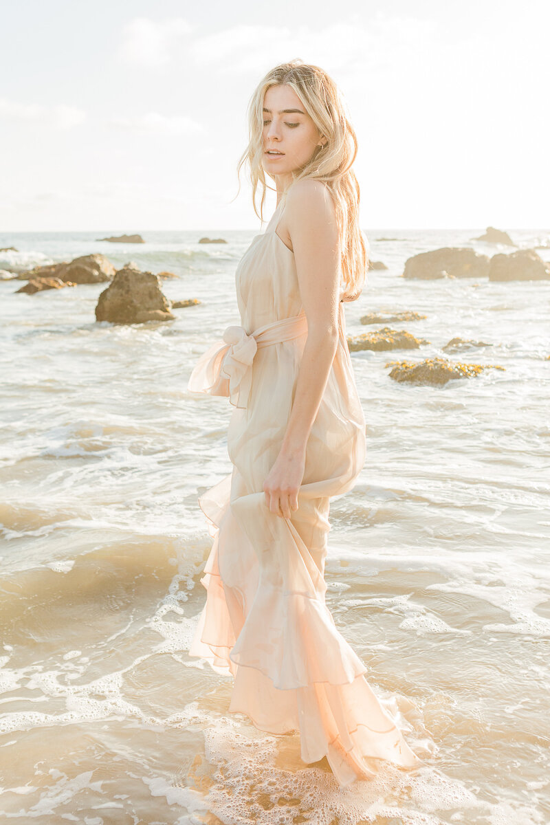 Top New England Wedding Photographer, Lia Rose Weddings, captures a bride standing in the ocean shoreline at sunset. The bottom of her dress is slightly wet. She is looking over her shoulder and down toward the water for a beautiful editorial-style bridal portrait.