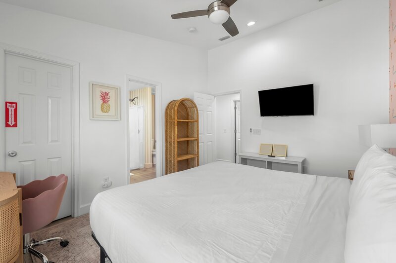Primary bedroom at the Surf House with smart TV, pink pineapple art, and access to en suite bathroom with vintage rattan shelf.