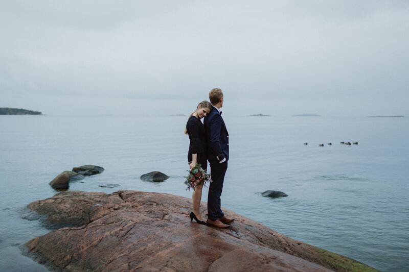 Morsian nojaa päätään sulhasen olkapäähän silmät kiinni ja sulhanen katsoo meressä uivia sorsia rantakallioilla Lauttasaaressa Helsingissä