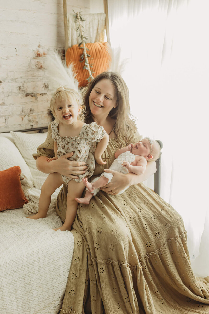 mom and toddler daughter on a bed together mom is also holding newborn baby sister and smiling in sibling newborn photos