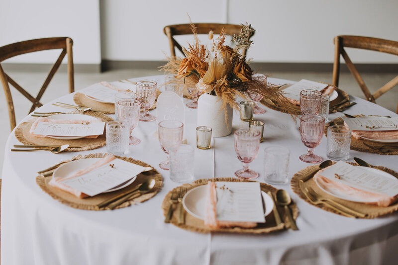Round wedding reception table styled with the Boho Dream decor package, featuring woven placemats, blush napkins, pink glassware, dried floral centerpieces, and gold accents for a soft bohemian look in Banff.