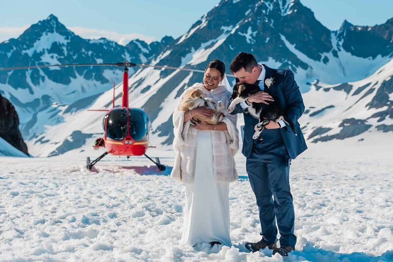 A bride and groom hold sled dog puppies after a dog sled across the glacier during their two day Bear Lake Glacier glamping adventure luxe elopement.  The groom holds a fluffy black and white dogsled puppy and kisses it on its head.  The bride is smiling broadly and laughing and holding a fluffy white and tan husky puppy.  The private helicopter they flew in on is bright red and viewable behind them, and the snow gleams white against the dark snowy mountains rising up from the glacier.