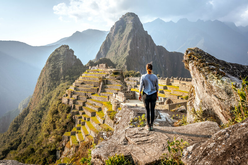 A person stands on a rocky ledge overlooking the ancient Incan city of Machu Picchu. The terraced ruins and surrounding mountains are bathed in soft sunlight under a partly cloudy sky.