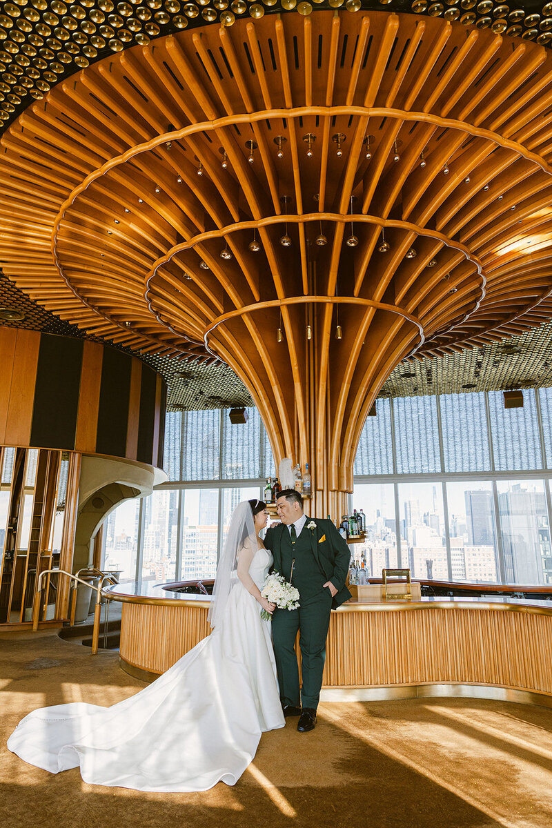 A bride and groom share a moment in an elegant loft space with floor-to-ceiling windows overlooking the city skyline in New York, NY.