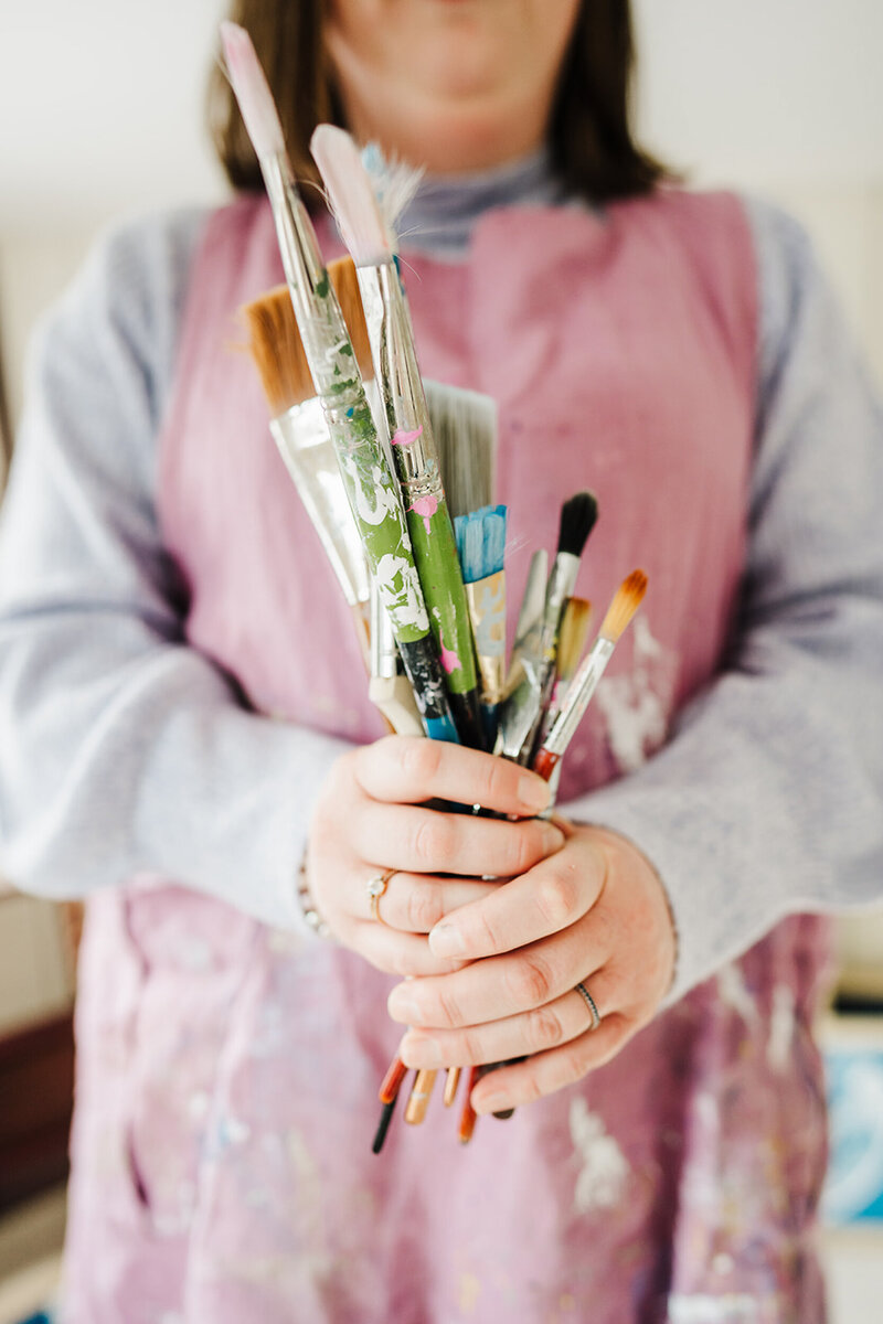 artist in pink smock holds paintbrushes for artist headshot