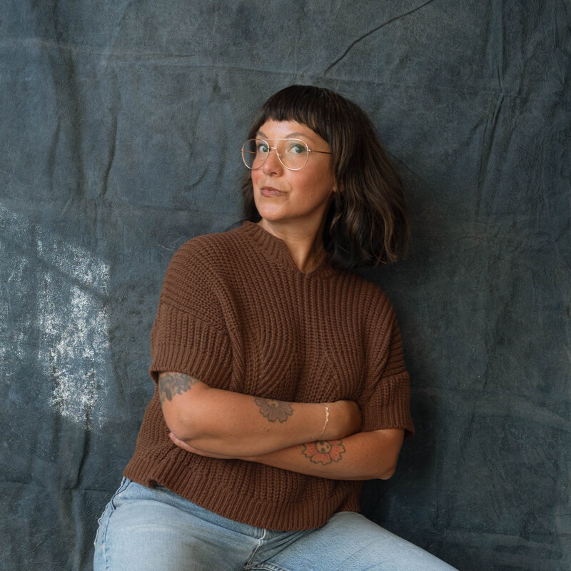 Maria Alcantara, Orange County family photographer, seated against a textured blue backdrop wearing a brown knit sweater and glasses.