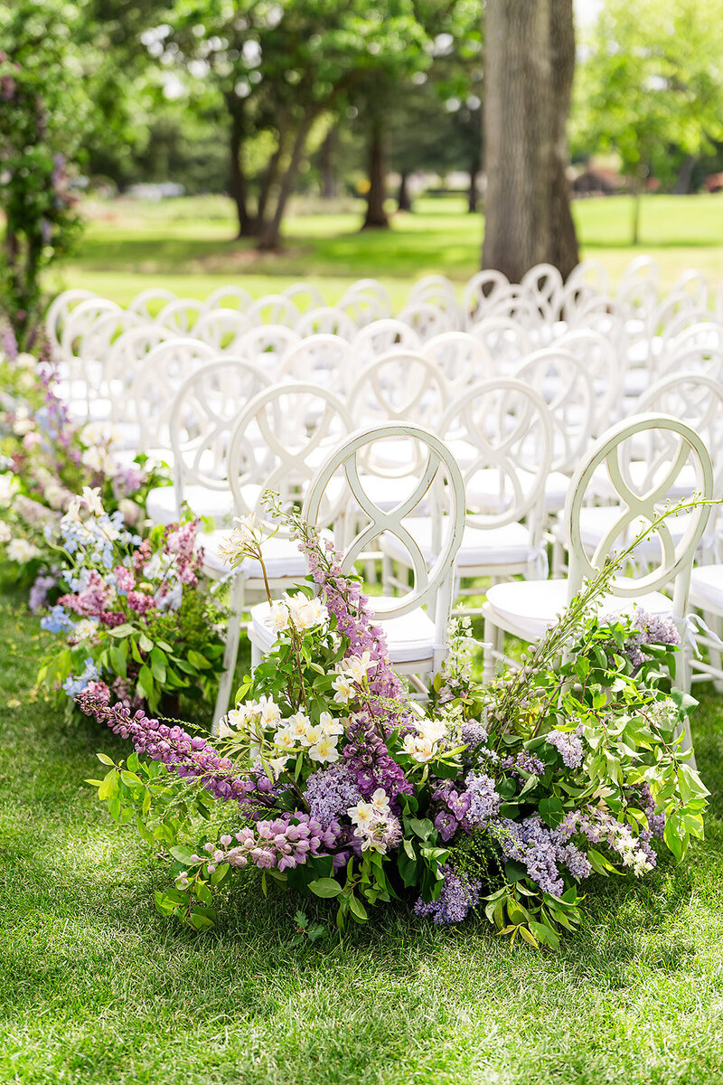 Ornate white chairs are arranged in rows on the grass outdoors with flowers in shades of purple and white
