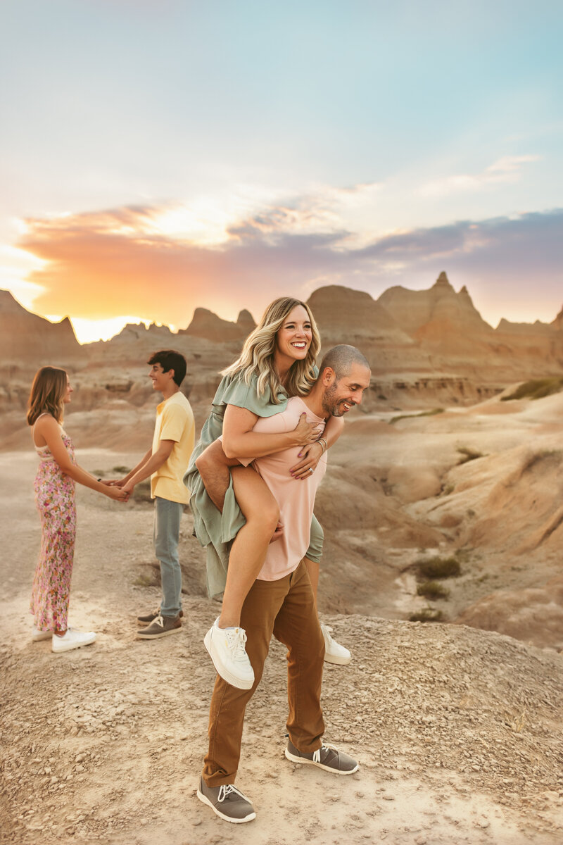 Mom gets piggy back ride on husband while kids play in background during photoshoot with Maternity Kansas City Photographers.