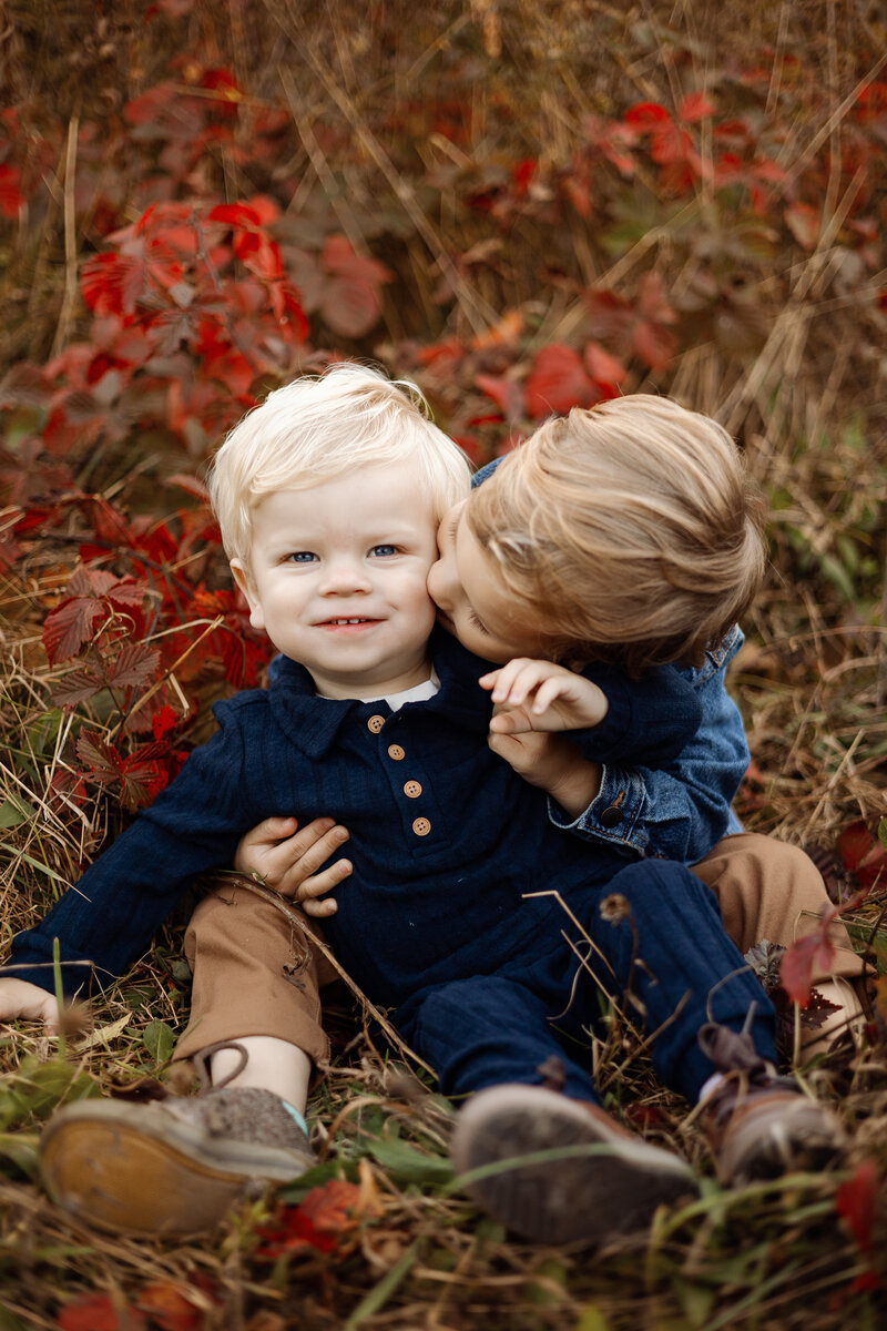 Two young brothers sitting in a field with autumn leaves in West Michigan, capturing a sweet and candid moment as one kisses the other's cheek during a seasonal family photography session.blanket. Captured by Honeywild Photography, a Grand Rapids, MI, luxury newborn photographer specializing in timeless and elegant portraits