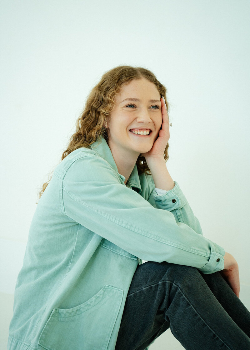 Haylee sitting on the ground with her elbow on her knee, holding her face. She is smiling and looking off to the side.